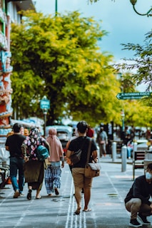 A person wearing a portable air quality monitor while walking through a tree-lined city street on a sunny day.