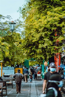 People are walking and interacting on a pathway lined with trees. Some individuals are seen from behind, while others are facing the camera. The area appears busy with several benches, a street lamp, and market stalls visible. The image has a lively urban atmosphere with lush green trees providing shade.