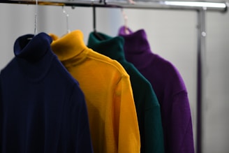 Four turtleneck sweaters in different colors hanging on a clothes rack. The sweaters are navy blue, yellow, green, and purple.