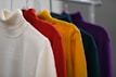 A row of colorful turtleneck sweaters displayed on hangers. The sweaters are arranged in a gradient of colors starting from white, then red, followed by yellow, green, and ending with purple. The soft texture of the fabric is noticeable, and the sweaters are neatly aligned on a clothing rack.