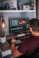 A person is sitting at a desk working on video editing software displayed on a large monitor. The desk is organized with decorative items like a globe, a plant, and a motivational sign saying 'Dream Big'. Camera lenses and other equipment are arranged on a shelf above. The person is holding a stylus and appears to be focused on their task.