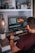 A person is sitting at a desk working on video editing software displayed on a large monitor. The desk is organized with decorative items like a globe, a plant, and a motivational sign saying 'Dream Big'. Camera lenses and other equipment are arranged on a shelf above. The person is holding a stylus and appears to be focused on their task.