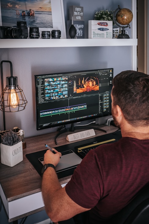 A person is sitting at a desk working on video editing software displayed on a large monitor. The desk is organized with decorative items like a globe, a plant, and a motivational sign saying 'Dream Big'. Camera lenses and other equipment are arranged on a shelf above. The person is holding a stylus and appears to be focused on their task.