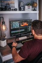 A person is sitting at a desk working on video editing software displayed on a large monitor. The desk is organized with decorative items like a globe, a plant, and a motivational sign saying 'Dream Big'. Camera lenses and other equipment are arranged on a shelf above. The person is holding a stylus and appears to be focused on their task.