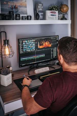 A person is sitting at a desk working on video editing software displayed on a large monitor. The desk is organized with decorative items like a globe, a plant, and a motivational sign saying 'Dream Big'. Camera lenses and other equipment are arranged on a shelf above. The person is holding a stylus and appears to be focused on their task.
