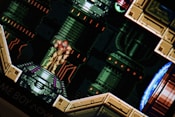 A pixelated scene from a video game featuring a character in a futuristic suit standing on a platform. The background consists of industrial, mechanical designs with pipes and panels.