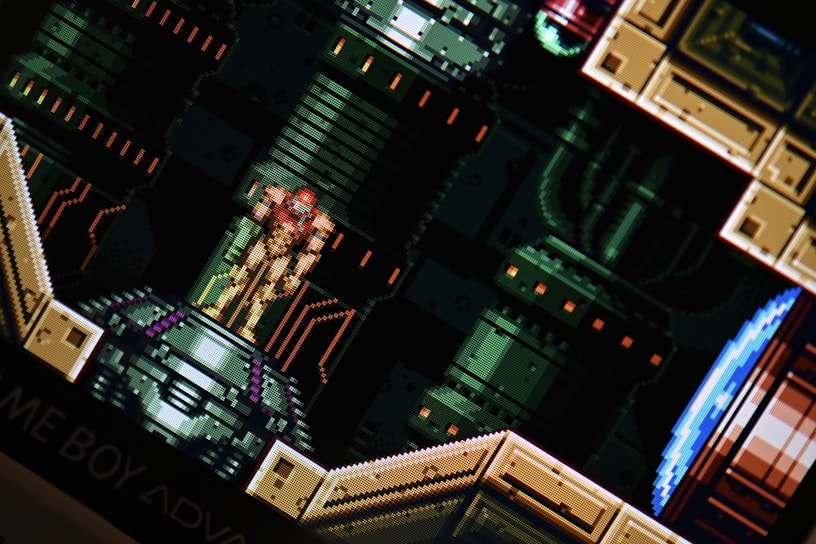 A pixelated scene from a video game featuring a character in a futuristic suit standing on a platform. The background consists of industrial, mechanical designs with pipes and panels.