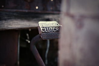 A close-up view of a metal clutch pedal with a textured grip. The word 'CLUTCH' is embossed on the surface of the pedal, which appears to be part of a rustic or vintage vehicle. The background is blurred, giving the focus entirely to the pedal.