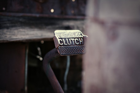A close-up view of a metal clutch pedal with a textured grip. The word 'CLUTCH' is embossed on the surface of the pedal, which appears to be part of a rustic or vintage vehicle. The background is blurred, giving the focus entirely to the pedal.