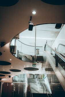 A modern interior space featuring an escalator, a flight of stairs, and a uniquely designed ceiling with circular cutouts and recessed lighting. The area is illuminated with soft lighting, creating an inviting atmosphere. The design incorporates reflective surfaces and clean lines, emphasizing a futuristic architectural style.