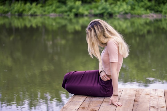 A person with long blonde hair sits on a wooden platform near a calm lake, surrounded by lush green foliage. Their posture is relaxed, suggesting a moment of peaceful contemplation.