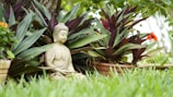 A serene stone statue of a meditating figure is surrounded by lush green foliage and purple-tinted leaves. The figure is sitting cross-legged, partially hidden by the dense vegetation. The scene conveys a sense of tranquility and natural harmony, enhanced by the bright green grass in the foreground.