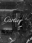 A store facade made of dark marble with prominent white veins runs vertically and horizontally. The name 'Cartier' is inscribed in elegant white script across the center. The reflection in the polished surface includes elements of an interior space such as a wall and possible furniture.