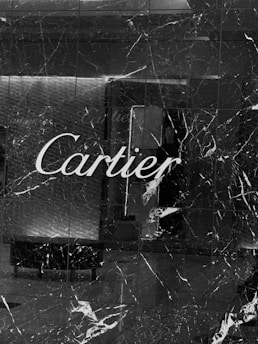 A store facade made of dark marble with prominent white veins runs vertically and horizontally. The name 'Cartier' is inscribed in elegant white script across the center. The reflection in the polished surface includes elements of an interior space such as a wall and possible furniture.