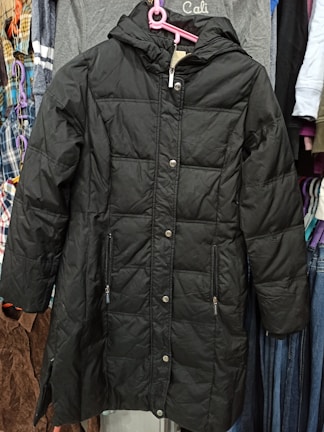 A black padded coat with a hood is displayed on a pink hanger. It features a quilted pattern with buttons down the front and two zippered pockets. The coat is hanging against a backdrop of various other clothing items, including shirts and jackets in different colors and patterns.