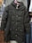 A black padded coat with a hood is displayed on a pink hanger. It features a quilted pattern with buttons down the front and two zippered pockets. The coat is hanging against a backdrop of various other clothing items, including shirts and jackets in different colors and patterns.