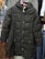 A black padded coat with a hood is displayed on a pink hanger. It features a quilted pattern with buttons down the front and two zippered pockets. The coat is hanging against a backdrop of various other clothing items, including shirts and jackets in different colors and patterns.