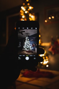A hand holding a smartphone capturing a photograph of a Christmas tree decorated with glowing lights and ornaments. The background is dimly lit, enhancing the warm glow of the lights on the tree.