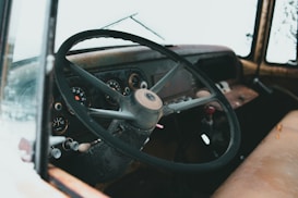 A vintage car interior showcasing an old steering wheel, a cluster of analog gauges on the dashboard, and a worn leather seat. The interior features a rustic, faded look with visible signs of age and wear.