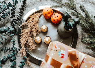 A festive arrangement featuring two clove-studded oranges and three wrapped chocolates on a silver tray. Surrounding the tray are sprigs of eucalyptus and a small evergreen branch. A vase with more foliage and a wrapped gift with a brown ribbon complete the scene.