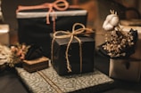 A collection of neatly wrapped gifts, featuring various textures and designs, some adorned with twine. One gift is decorated with a small bouquet of cotton and dried flowers, adding a rustic and elegant touch.