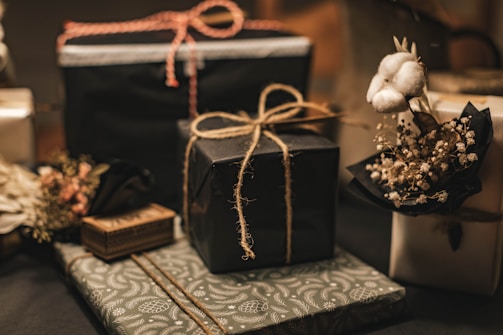 A collection of neatly wrapped gifts, featuring various textures and designs, some adorned with twine. One gift is decorated with a small bouquet of cotton and dried flowers, adding a rustic and elegant touch.
