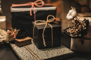 A collection of neatly wrapped gifts, featuring various textures and designs, some adorned with twine. One gift is decorated with a small bouquet of cotton and dried flowers, adding a rustic and elegant touch.