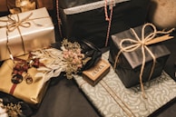 Multiple gift boxes are wrapped in elegant paper with a variety of colors and textures. The packages feature intricate patterns and some are tied with simple twine. A small dried floral arrangement adds a touch of rustic charm to one of the gifts.