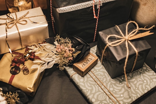 Multiple gift boxes are wrapped in elegant paper with a variety of colors and textures. The packages feature intricate patterns and some are tied with simple twine. A small dried floral arrangement adds a touch of rustic charm to one of the gifts.