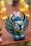 A colorful, intricately designed figurine with fierce facial features and elaborate wings. The sculpture includes bright blue, green, and yellow accents with bold black horns and detailed facial expressions. It stands on a reddish-brown surface with blurred yellow figures in the background.