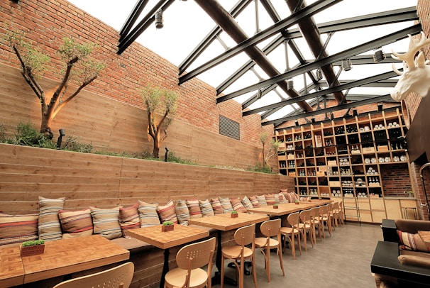 A warmly lit, stylish restaurant interior featuring wooden tables and chairs lined up along a long bench seat that is adorned with colorful pillows. The space has a modern, industrial aesthetic with exposed brick walls and a large shelf filled with wine bottles and jars. The ceiling is made of glass and steel beams, allowing natural light to illuminate the room, and plant arrangements add a touch of greenery.