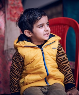A young child is sitting on a bright red plastic chair. The child is wearing a yellow puffer vest with a blue zipper over a patterned long-sleeve shirt. The background appears to be indoors, with partially visible items including fabric with red and beige tones.