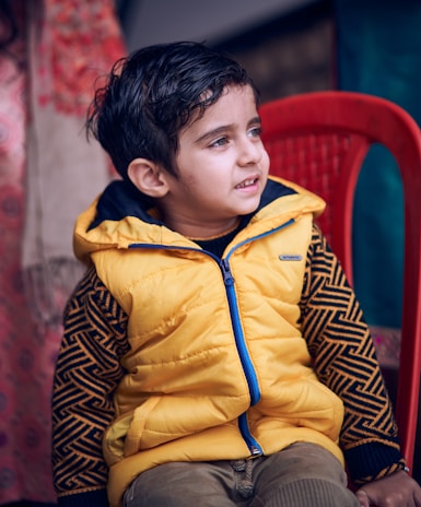 A young child is sitting on a bright red plastic chair. The child is wearing a yellow puffer vest with a blue zipper over a patterned long-sleeve shirt. The background appears to be indoors, with partially visible items including fabric with red and beige tones.