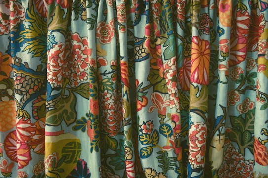 A close-up view of a vibrant, intricately patterned fabric, featuring an array of colorful floral designs. The rich details include various blooming flowers in shades of red, pink, and yellow, interspersed with green leaves and blue accents. The fabric appears gathered, creating a textured surface with folds and shadows.