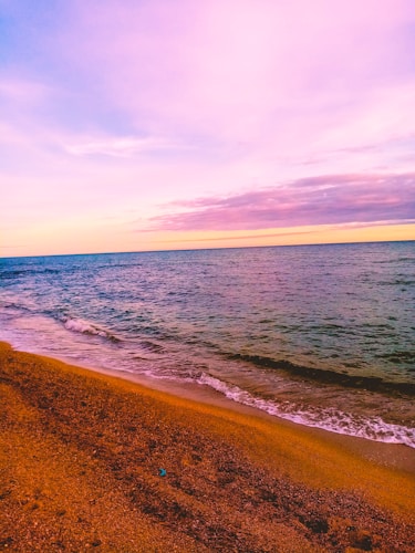 A serene beach scene with gentle waves lapping against a sandy shore. The sky is filled with vibrant pastel colors, primarily shades of pink and purple, suggesting a sunset or sunrise.