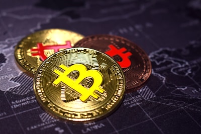 Three Bitcoin coins with the iconic 'B' logo rest on a dark world map. The coins display a shiny, golden and copper metallic finish, suggesting value and currency.