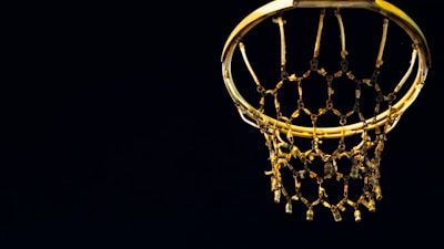 A close-up view of a basketball hoop and net dramatically lit against a dark background. The metallic hoop and the net are visible, with the net showing chain-like elements.