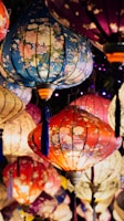 Colorful lanterns with floral patterns hang together, creating a vibrant and festive display. The lanterns vary in color from blue and red to purple and beige, each adorned with delicate floral designs that enhance their beauty. The warm glow from the lanterns adds to the festive and joyful atmosphere.