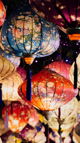 Colorful lanterns with floral patterns hang together, creating a vibrant and festive display. The lanterns vary in color from blue and red to purple and beige, each adorned with delicate floral designs that enhance their beauty. The warm glow from the lanterns adds to the festive and joyful atmosphere.