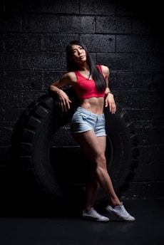 A woman is leaning against a large tire in a dimly lit gym environment. She is wearing a red sports bra, denim shorts, and white sneakers. Her pose is relaxed and confident, emphasizing her athletic build.