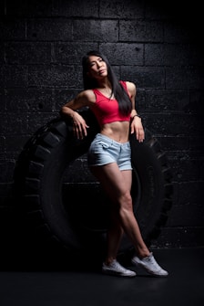 A woman is leaning against a large tire in a dimly lit gym environment. She is wearing a red sports bra, denim shorts, and white sneakers. Her pose is relaxed and confident, emphasizing her athletic build.