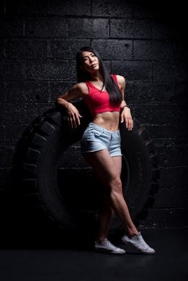 A woman is leaning against a large tire in a dimly lit gym environment. She is wearing a red sports bra, denim shorts, and white sneakers. Her pose is relaxed and confident, emphasizing her athletic build.