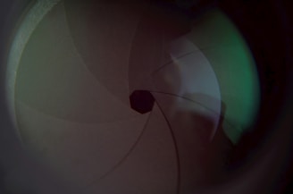 The image displays a close-up view of an opened camera lens diaphragm with overlapping blades creating a polygonal aperture shape in the center. The lighting casts gentle reflections and patterns on the surface, with subtle shades of green and brown visible.