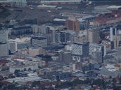 An aerial view of an urban landscape with numerous multi-story buildings, including office complexes and residential structures. The scene includes a mix of modern architecture, industrial areas, and residential neighborhoods. Roads and pathways are visible, along with a harbor area in the background.