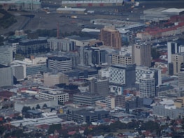 An aerial view of an urban landscape with numerous multi-story buildings, including office complexes and residential structures. The scene includes a mix of modern architecture, industrial areas, and residential neighborhoods. Roads and pathways are visible, along with a harbor area in the background.