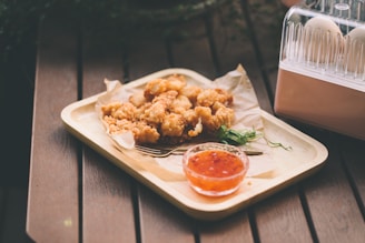 On a wooden surface, a wooden tray holds crispy fried chicken pieces lined with parchment paper. Next to the chicken, there is a small glass dish filled with a red dipping sauce. A few sprigs of fresh herbs are placed for garnish. To the side, there is an egg organizer holding several eggs.