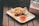 On a wooden surface, a wooden tray holds crispy fried chicken pieces lined with parchment paper. Next to the chicken, there is a small glass dish filled with a red dipping sauce. A few sprigs of fresh herbs are placed for garnish. To the side, there is an egg organizer holding several eggs.