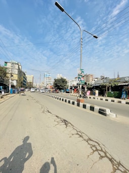 A wide street with cracked pavement running through an urban area, flanked by multiple-story buildings on both sides. Tall streetlights line the road, and overhead power lines are visible. A few vehicles, including rickshaws and motorbikes, are present along the road. There are shadows of bicyclists on the street, indicating the position of the sun.