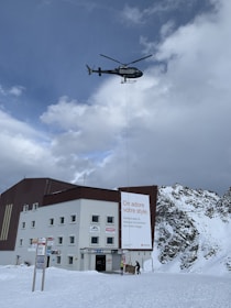 A helicopter is hovering in the sky with mountains in the background. Below, a building with advertisements is visible, and there is snow on the ground. People are present, possibly engaged in unloading cargo.