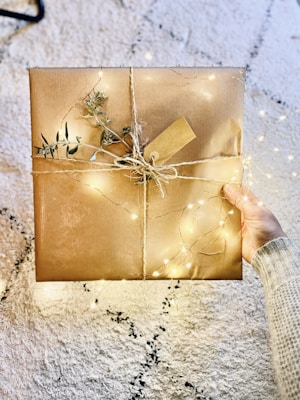 A hand holding a neatly wrapped gift box adorned with brown paper and twine. The package is embellished with small fairy lights and sprigs of greenery, giving it an elegant and festive appearance. It rests on a soft, textured rug with a neutral color palette.